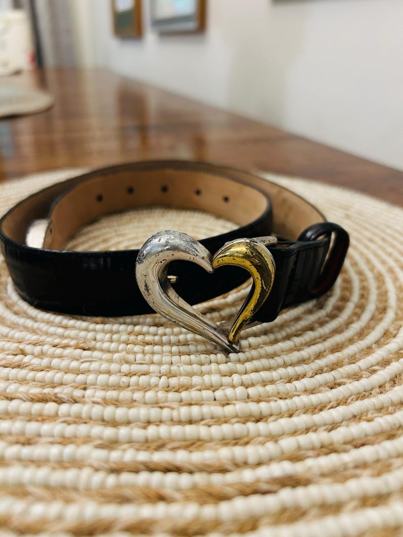 May include: Black belt with a silver and gold heart-shaped buckle. The belt is made of a textured material and has a classic design. The buckle is a prominent feature, adding a touch of elegance to the accessory.