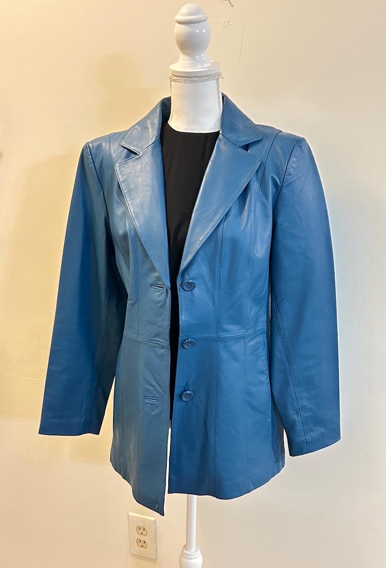 K&ouml;nnte beinhalten: Ein blauer Lederblazer mit klassischem Revers und Dreiknopfverschluss. Die Jacke wird auf einer Schaufensterpuppe pr&auml;sentiert, die ihre ma&szlig;geschneiderte Passform und ihr Design hervorhebt. Der Blazer hat ein schlankes, poliertes Aussehen.