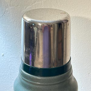 May include: A vacuum insulated thermos featuring a textured sage green body and a polished silver metallic cap. The cylindrical container has a dark green band separating the main body from the reflective metal lid, which shows a faint reflection.