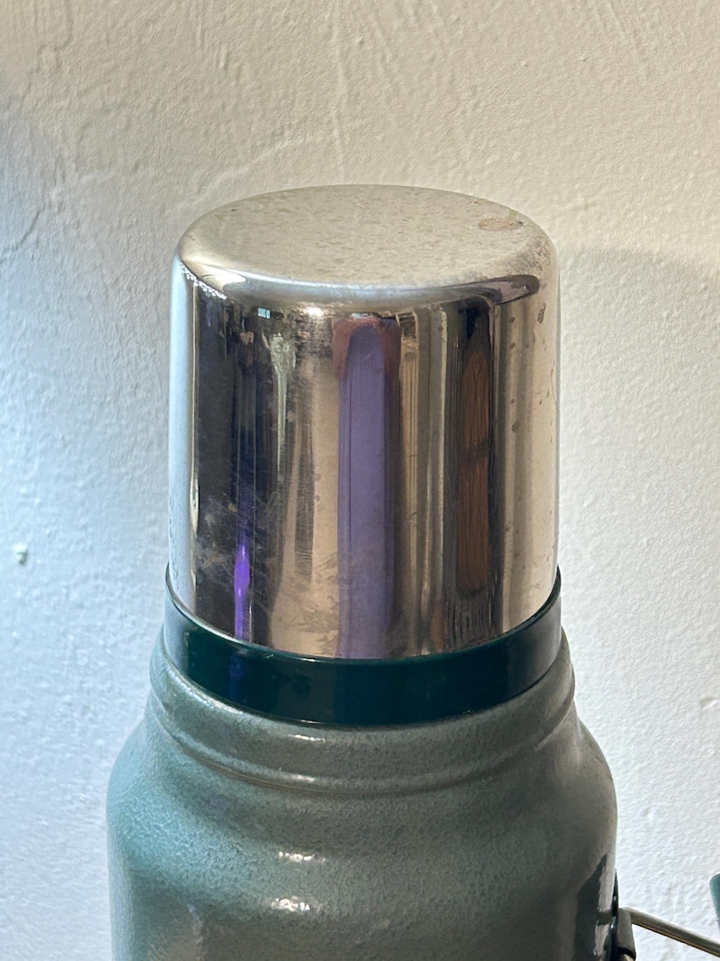 May include: A vacuum insulated thermos featuring a textured sage green body and a polished silver metallic cap. The cylindrical container has a dark green band separating the main body from the reflective metal lid, which shows a faint reflection.