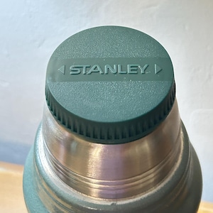 May include: A sage green vacuum insulated bottle featuring a matching green screw-top lid with a textured rim. The top of the lid displays the word STANLEY in embossed capital letters, flanked by two small triangular icons pointing inward.