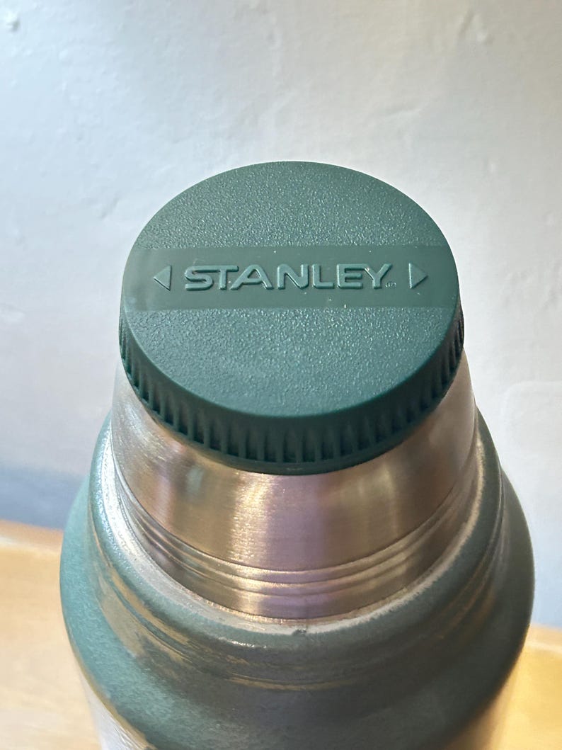 May include: A sage green vacuum insulated bottle featuring a matching green screw-top lid with a textured rim. The top of the lid displays the word STANLEY in embossed capital letters, flanked by two small triangular icons pointing inward.