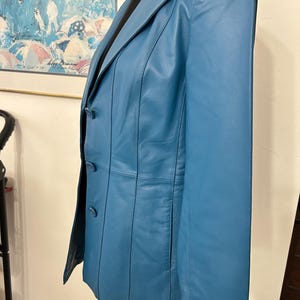 K&ouml;nnte beinhalten: Ein teal-farbener Lederblazer mit fallendem Revers und Dreiknopfverschluss. Die Jacke hat vertikale N&auml;hte und eine taillierte Passform. Der Blazer wird auf einer wei&szlig;en Schaufensterpuppe pr&auml;sentiert und hat ein schwarzes Futter.