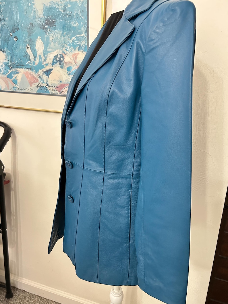K&ouml;nnte beinhalten: Ein teal-farbener Lederblazer mit fallendem Revers und Dreiknopfverschluss. Die Jacke hat vertikale N&auml;hte und eine taillierte Passform. Der Blazer wird auf einer wei&szlig;en Schaufensterpuppe pr&auml;sentiert und hat ein schwarzes Futter.