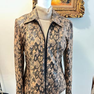 May include: A tan and black snakeskin-patterned jacket with a black zipper closure. The jacket is displayed on a mannequin with a beige turtleneck underneath. The jacket has a collared neckline and long sleeves.