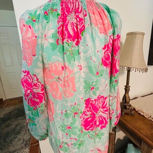 May include: A long-sleeved blouse with a floral design. The blouse has large pink flowers, green leaves, and white details on a light blue backdrop. The sleeves are long and gathered at the cuffs.