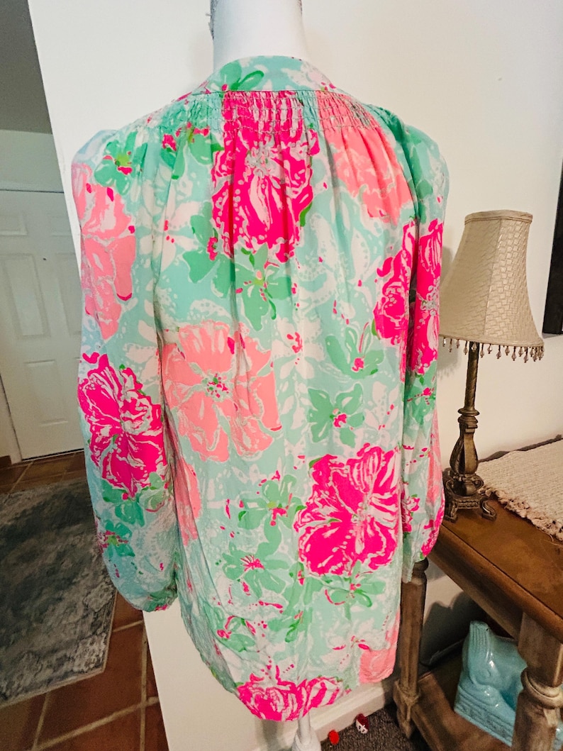 May include: A long-sleeved blouse with a floral design. The blouse has large pink flowers, green leaves, and white details on a light blue backdrop. The sleeves are long and gathered at the cuffs.