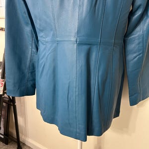 K&ouml;nnte beinhalten: T&uuml;rkisfarbene Lederjacke mit langen &Auml;rmeln. Die Jacke hat vertikale Steppn&auml;hte und eine taillierte Passform. Die R&uuml;ckseite der Jacke ist sichtbar und zeigt die N&auml;hte und das Design. Die Jacke wird auf einer wei&szlig;en Schaufensterpuppe pr&auml;sentiert.