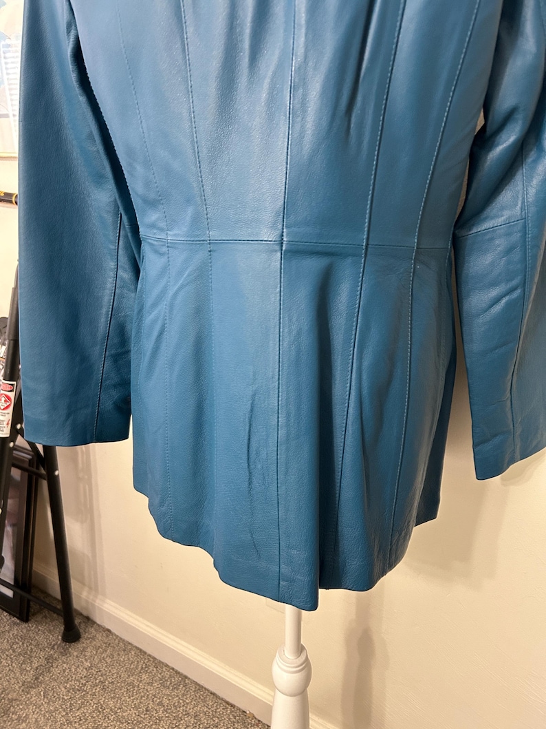 K&ouml;nnte beinhalten: T&uuml;rkisfarbene Lederjacke mit langen &Auml;rmeln. Die Jacke hat vertikale Steppn&auml;hte und eine taillierte Passform. Die R&uuml;ckseite der Jacke ist sichtbar und zeigt die N&auml;hte und das Design. Die Jacke wird auf einer wei&szlig;en Schaufensterpuppe pr&auml;sentiert.