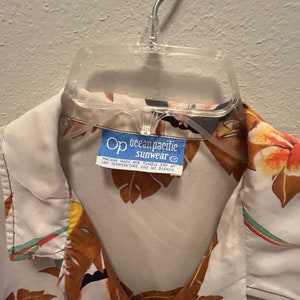May include: A white button-down shirt with a tropical print featuring brown leaves, colorful birds, and flowers. The shirt has a blue label that reads "Op ocean pacific sunwear" with care instructions.