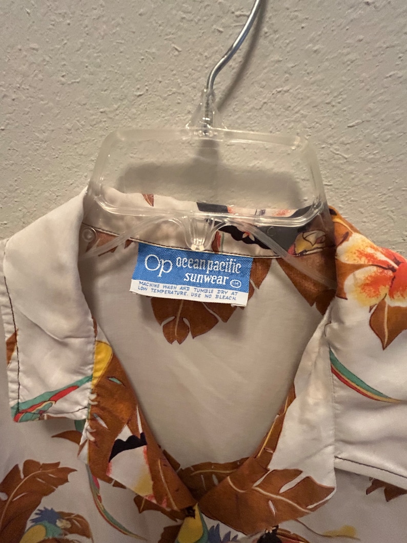 May include: A white button-down shirt with a tropical print featuring brown leaves, colorful birds, and flowers. The shirt has a blue label that reads "Op ocean pacific sunwear" with care instructions.