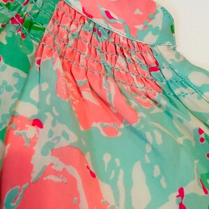May include: Close-up of a patterned blouse in pink, turquoise, and white abstract shapes. The neckline and upper bodice have a smocked detail. The fabric appears lightweight and flowy, suitable for warmer weather. The design is eye-catching.