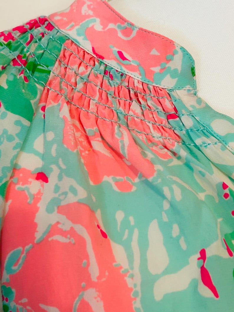 May include: Close-up of a patterned blouse in pink, turquoise, and white abstract shapes. The neckline and upper bodice have a smocked detail. The fabric appears lightweight and flowy, suitable for warmer weather. The design is eye-catching.