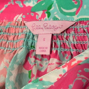 May include: Close-up of a colourful garment with a pink label that reads "Lilly Pulitzer". The garment features a vibrant floral pattern in shades of pink, turquoise, and green. The label indicates the size is "S" and "Made in China".
