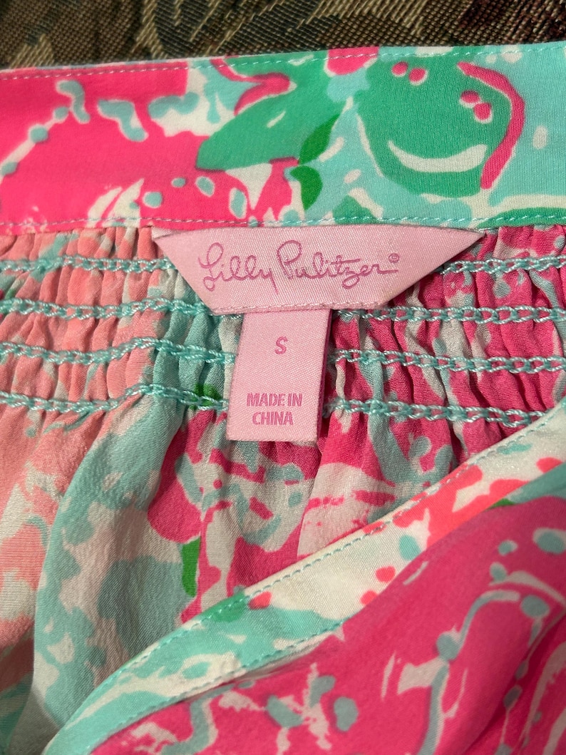 May include: Close-up of a colourful garment with a pink label that reads "Lilly Pulitzer". The garment features a vibrant floral pattern in shades of pink, turquoise, and green. The label indicates the size is "S" and "Made in China".