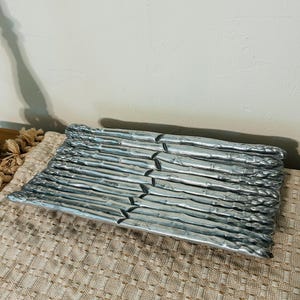 May include: A silver-colored, rectangular serving tray with a textured surface, shaped like asparagus spears. The tray is filled with multiple asparagus spears. The tray is resting on a beige woven placemat.