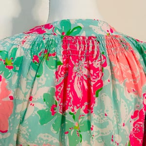 May include: A close-up of a vibrant floral print top. The fabric displays a pattern of large pink and green flowers on a light blue and white backdrop. The top has a gathered neckline and a loose fit.