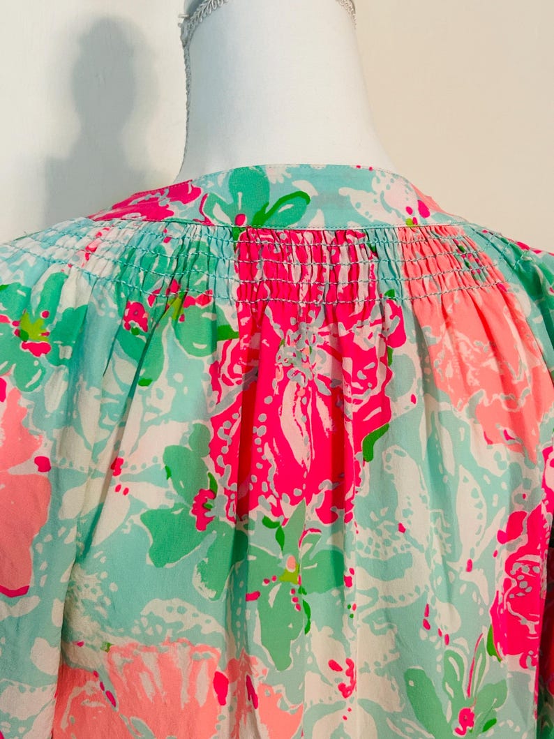 May include: A close-up of a vibrant floral print top. The fabric displays a pattern of large pink and green flowers on a light blue and white backdrop. The top has a gathered neckline and a loose fit.