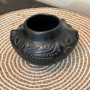 May include: A black ceramic bowl with a wide opening and a rounded base. The bowl features embossed patterns of circles and curved lines. It sits on a round woven placemat with alternating beige and white concentric circles.