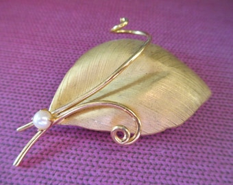 May include: Gold-toned leaf-shaped brooch with a textured surface. The brooch features a delicate, curved stem and a small, round pearl accent. The design includes a curled accent at the top. The brooch is a vintage-style jewelry piece.