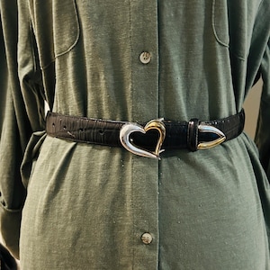 May include: Black belt with a silver and gold heart-shaped buckle. The belt has a textured, crocodile-like pattern. The buckle is a prominent feature, adding a decorative element to the belt. The belt is worn over a green dress.