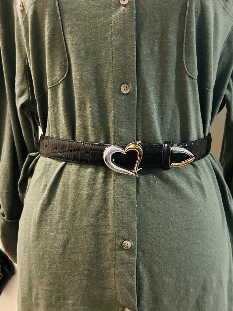 May include: Black belt with a silver and gold heart-shaped buckle. The belt has a textured, crocodile-like pattern. The buckle is a prominent feature, adding a decorative element to the belt. The belt is worn over a green dress.