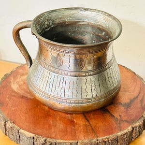 May include: An antique copper mug with a handle, featuring etched designs and a tarnished finish. The mug has a wide mouth and a bulbous body, resting on a wooden slice.