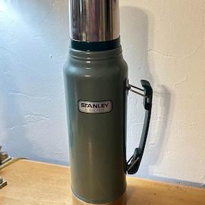 May include: A tall, cylindrical green vacuum-insulated flask with a metallic silver cap and base. The body features a dark green plastic handle attached to the side and a white rectangular label with the black text STANLEY SINCE 1913.