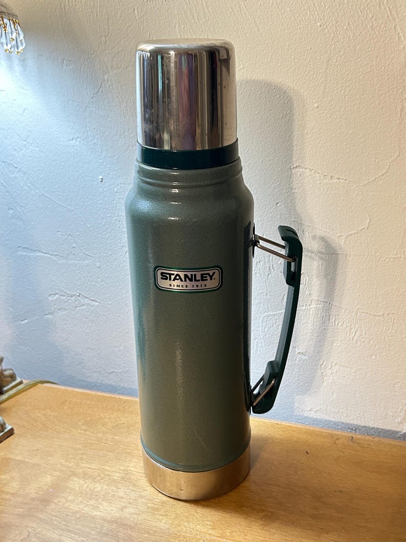 May include: A tall, cylindrical green vacuum-insulated flask with a metallic silver cap and base. The body features a dark green plastic handle attached to the side and a white rectangular label with the black text STANLEY SINCE 1913.