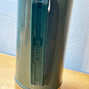 May include: A dark green cylindrical vacuum insulated bottle with a metallic base and a dark green plastic handle. The handle features the word STANLEY embossed vertically in capital letters. The bottle has a textured finish and shows minor wear.