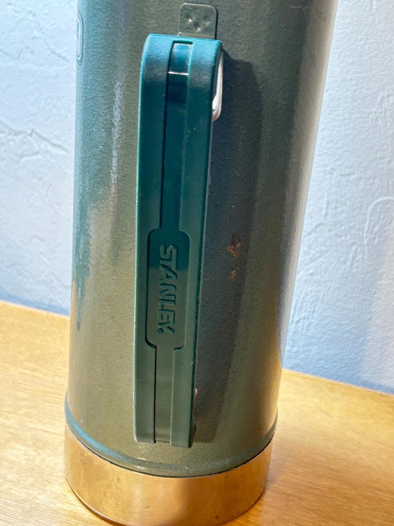 May include: A dark green cylindrical vacuum insulated bottle with a metallic base and a dark green plastic handle. The handle features the word STANLEY embossed vertically in capital letters. The bottle has a textured finish and shows minor wear.