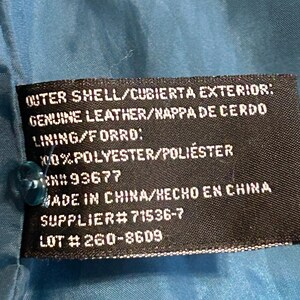 K&ouml;nnte beinhalten: Nahaufnahme eines schwarzen Etiketts mit wei&szlig;em Text, befestigt an einem t&uuml;rkisfarbenen Stoff. Das Etikett gibt die Au&szlig;enschicht als echtes Leder, das Futter als Polyester an und enth&auml;lt eine Lieferantennummer und Losnummer. Das Etikett besagt auch "Made in China".