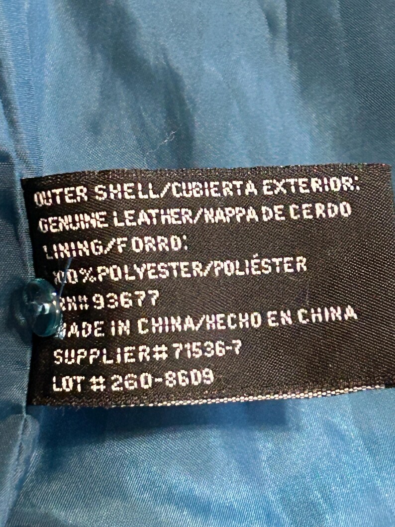 K&ouml;nnte beinhalten: Nahaufnahme eines schwarzen Etiketts mit wei&szlig;em Text, befestigt an einem t&uuml;rkisfarbenen Stoff. Das Etikett gibt die Au&szlig;enschicht als echtes Leder, das Futter als Polyester an und enth&auml;lt eine Lieferantennummer und Losnummer. Das Etikett besagt auch "Made in China".