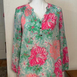 May include: A long-sleeved blouse with a floral design. The blouse has a V-neck and is decorated with large pink and smaller green flowers on a light blue backdrop. The sleeves are long and gathered at the cuffs. The blouse is displayed on a mannequin.