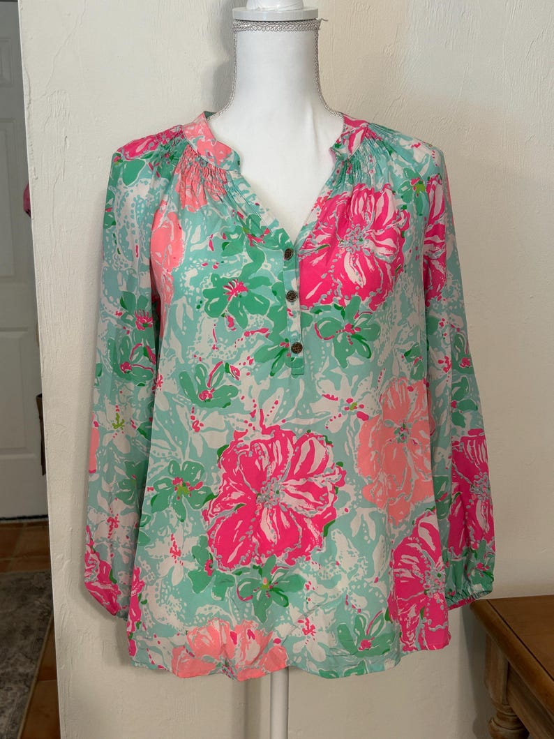 May include: A long-sleeved blouse with a floral design. The blouse has a V-neck and is decorated with large pink and smaller green flowers on a light blue backdrop. The sleeves are long and gathered at the cuffs. The blouse is displayed on a mannequin.