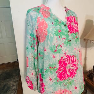 May include: A long-sleeved blouse with a floral pattern. The blouse has a light blue background with large pink and green floral designs. It features a V-neckline with three buttons and gathered detailing at the shoulders.