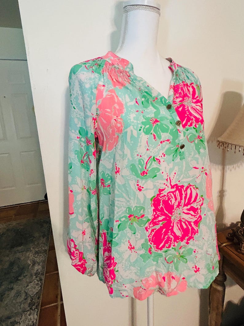 May include: A long-sleeved blouse with a floral pattern. The blouse has a light blue background with large pink and green floral designs. It features a V-neckline with three buttons and gathered detailing at the shoulders.