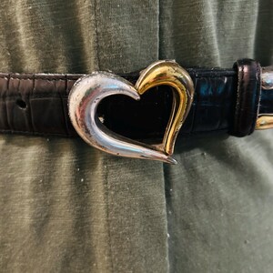May include: Black belt with a silver and gold heart-shaped buckle. The belt has a textured, crocodile-like pattern. The heart buckle is split, with one side silver and the other gold.