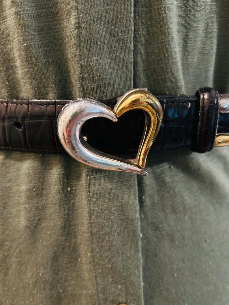 May include: Black belt with a silver and gold heart-shaped buckle. The belt has a textured, crocodile-like pattern. The heart buckle is split, with one side silver and the other gold.