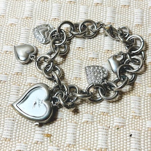 May include: Silver-toned bracelet with heart-shaped charms and a heart-shaped watch face. The watch face has a white background and black hands. The bracelet has a chain link design and several heart-shaped charms.