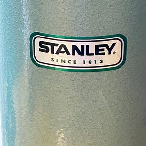 May include: A light blue, textured cylindrical container featuring a rectangular logo with rounded corners. The logo displays the word STANLEY in bold black letters above the text SINCE 1913, all enclosed within a dark green border.