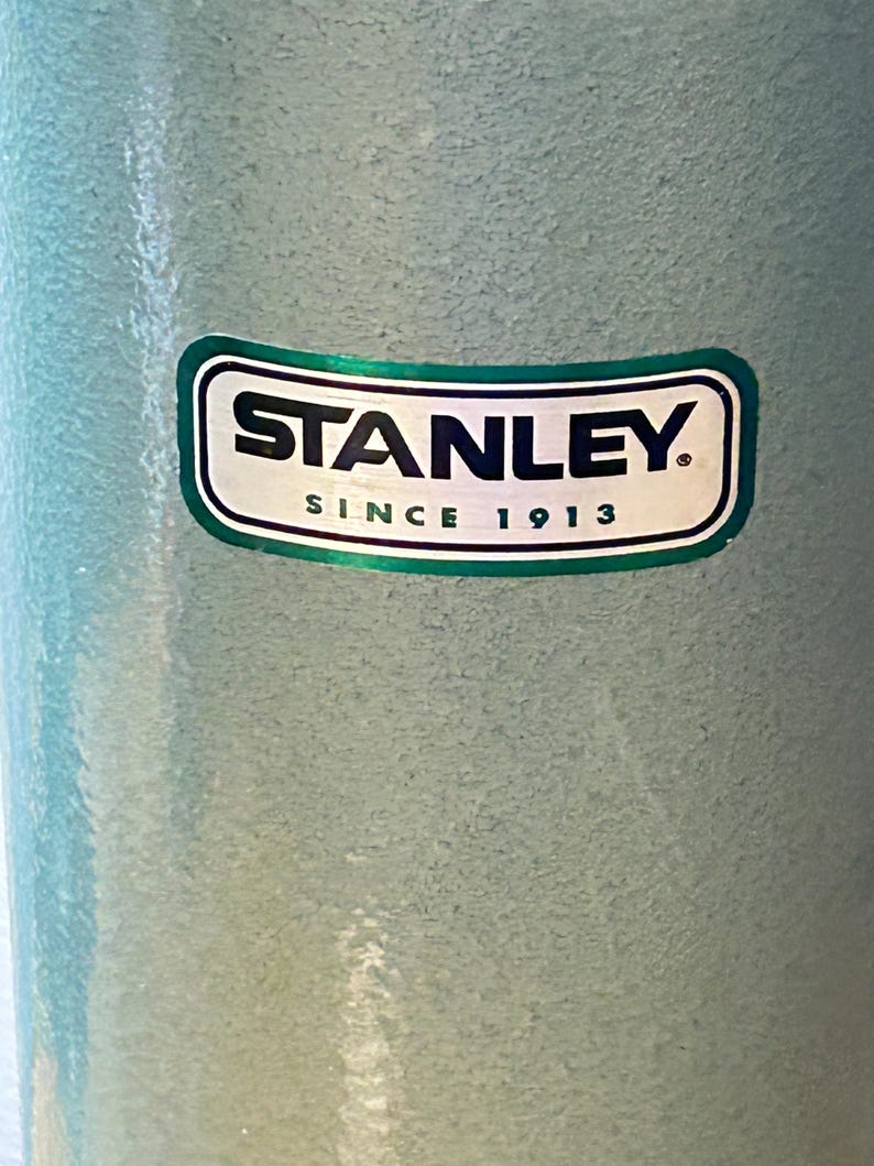 May include: A light blue, textured cylindrical container featuring a rectangular logo with rounded corners. The logo displays the word STANLEY in bold black letters above the text SINCE 1913, all enclosed within a dark green border.