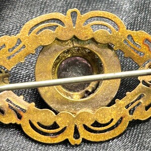 Op de afbeelding: Vintage goudkleurige broche met een sierlijk, opengewerkt ontwerp. De broche heeft een centrale, cirkelvormige zetting met een paarse edelsteen. De speld is bevestigd met een eenvoudige sluiting aan de achterkant.