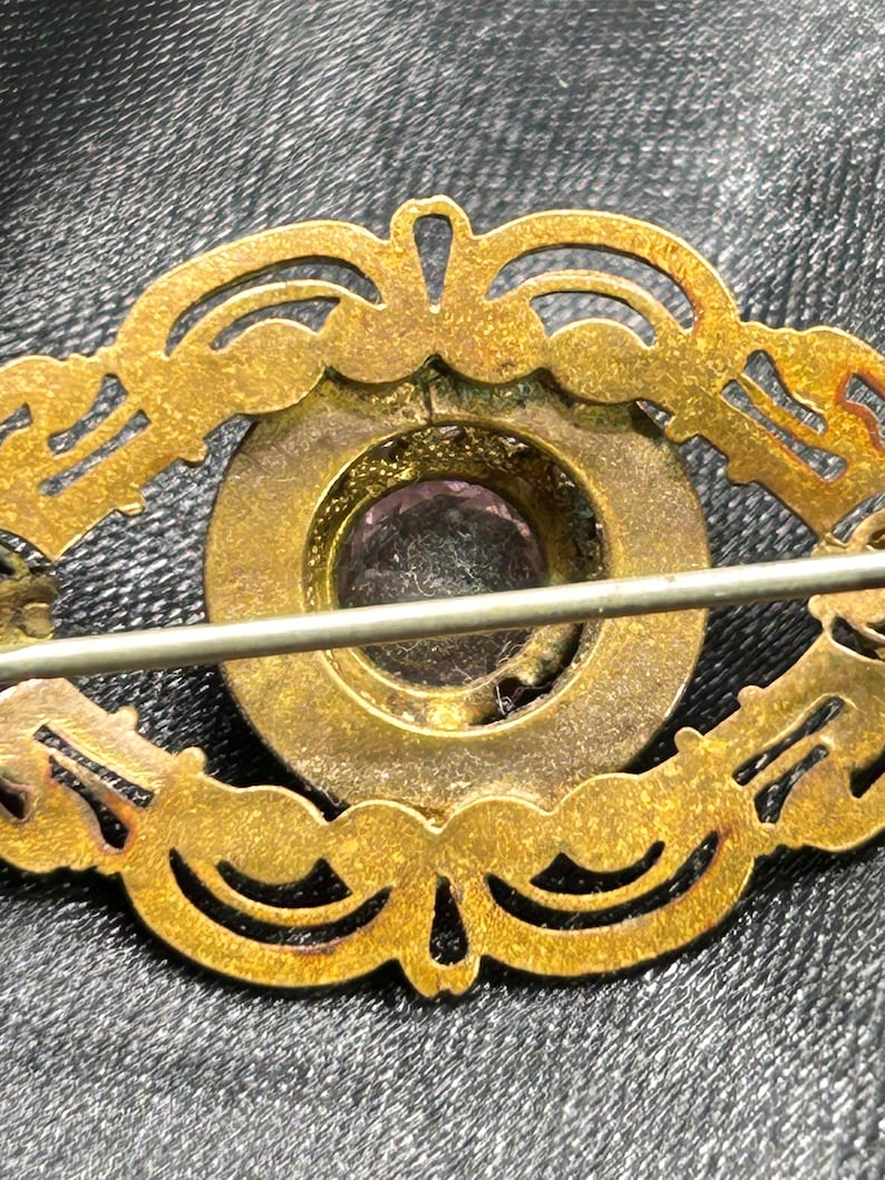 Op de afbeelding: Vintage goudkleurige broche met een sierlijk, opengewerkt ontwerp. De broche heeft een centrale, cirkelvormige zetting met een paarse edelsteen. De speld is bevestigd met een eenvoudige sluiting aan de achterkant.