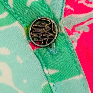May include: Close-up of a button on patterned fabric. The silver-toned button has an ornate design. The fabric displays a vibrant pattern of green, white, and pink, with a turquoise border.