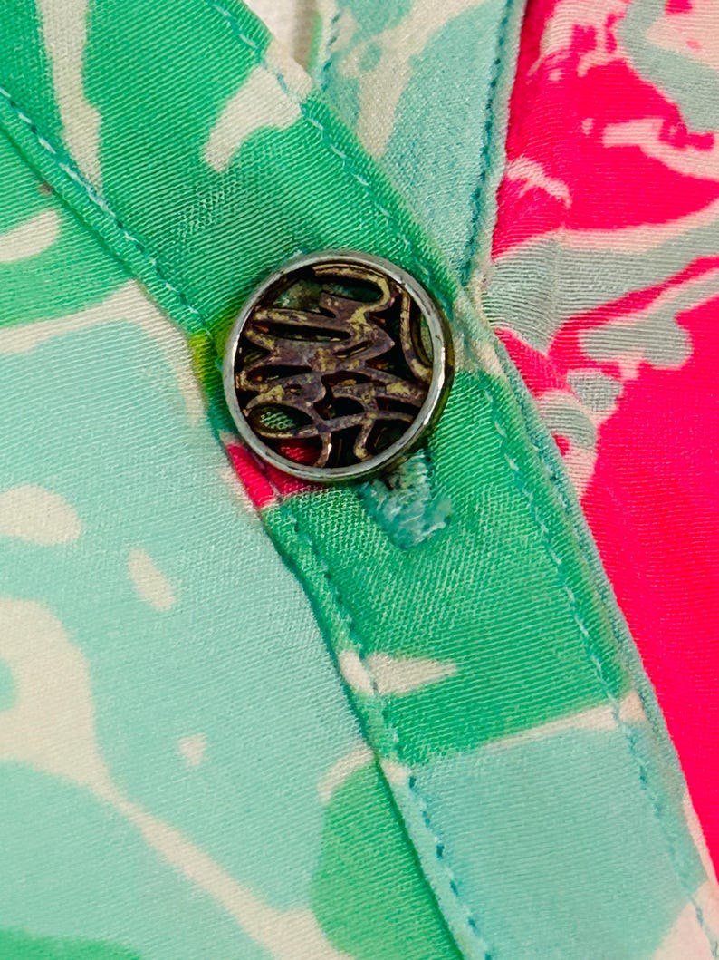 May include: Close-up of a button on patterned fabric. The silver-toned button has an ornate design. The fabric displays a vibrant pattern of green, white, and pink, with a turquoise border.