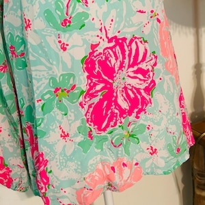 May include: A sleeveless top with a vibrant floral pattern. The fabric features large, bright pink flowers, smaller green and pink blooms, and white accents on a light blue background. The top has a buttoned neckline.