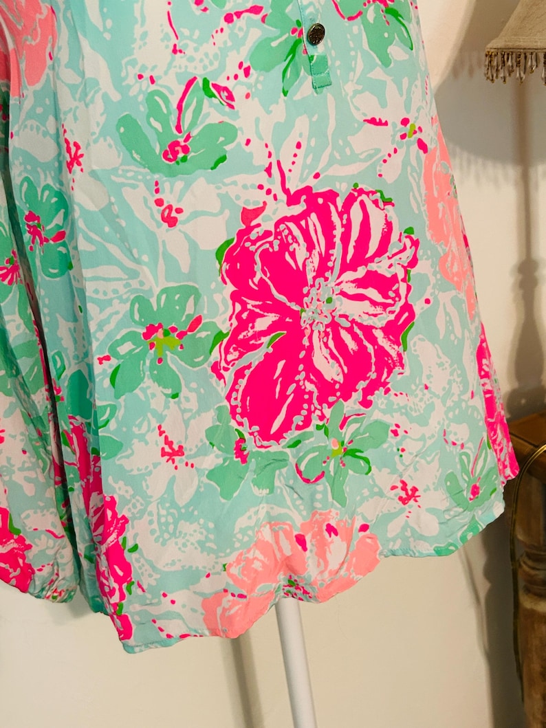 May include: A sleeveless top with a vibrant floral pattern. The fabric features large, bright pink flowers, smaller green and pink blooms, and white accents on a light blue background. The top has a buttoned neckline.