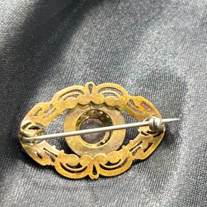 Op de afbeelding: Ovale antieke broche met een goudkleurig filigraan ontwerp. Het midden heeft een donkerpaarse steen omgeven door een goudkleurige metalen zetting. De broche heeft een speldsluiting aan de achterkant.