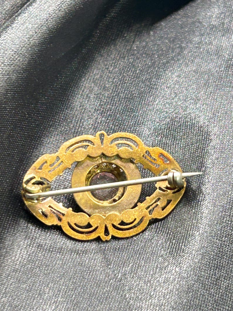 Op de afbeelding: Ovale antieke broche met een goudkleurig filigraan ontwerp. Het midden heeft een donkerpaarse steen omgeven door een goudkleurige metalen zetting. De broche heeft een speldsluiting aan de achterkant.
